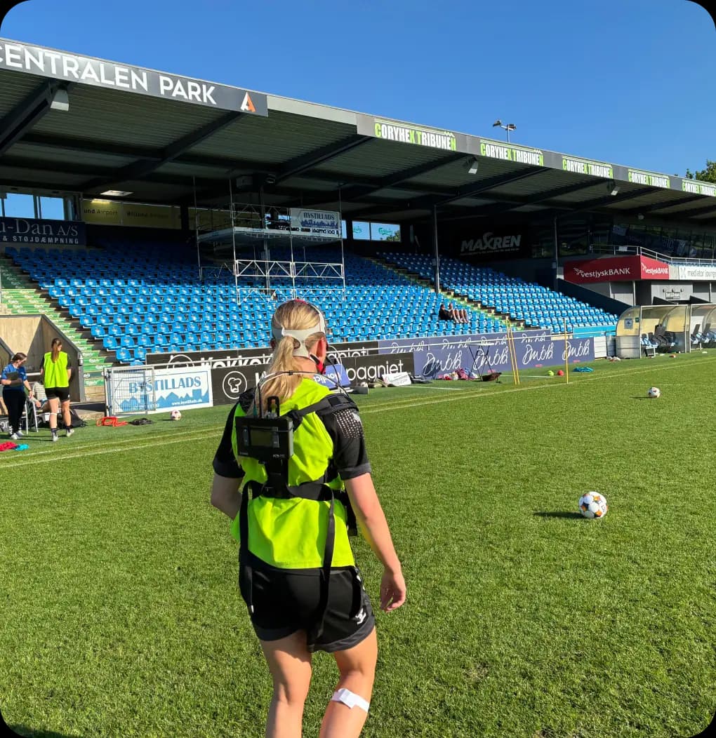 Female football player wearing a GPS vest during a training session at a stadium with blue seats and bright sunlight.