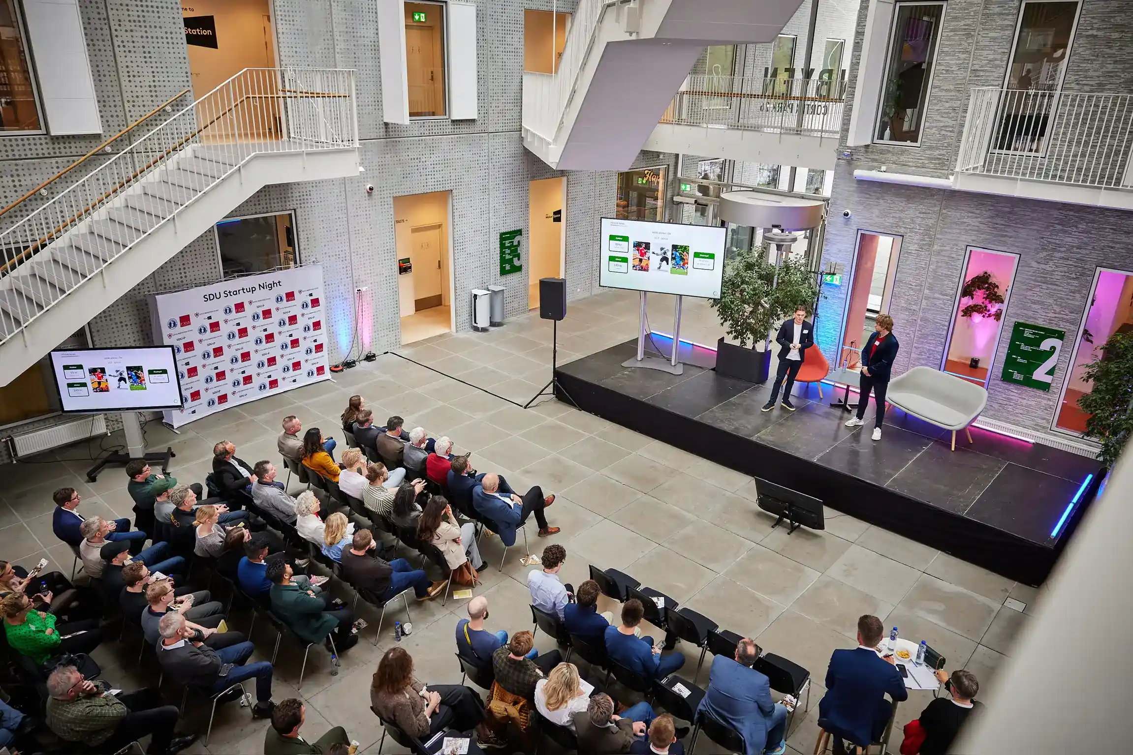 Presentation at SDU Startup Night held in a modern atrium. Two speakers stand on stage in front of a seated audience, with large screens displaying slides and a backdrop featuring sponsor logos.